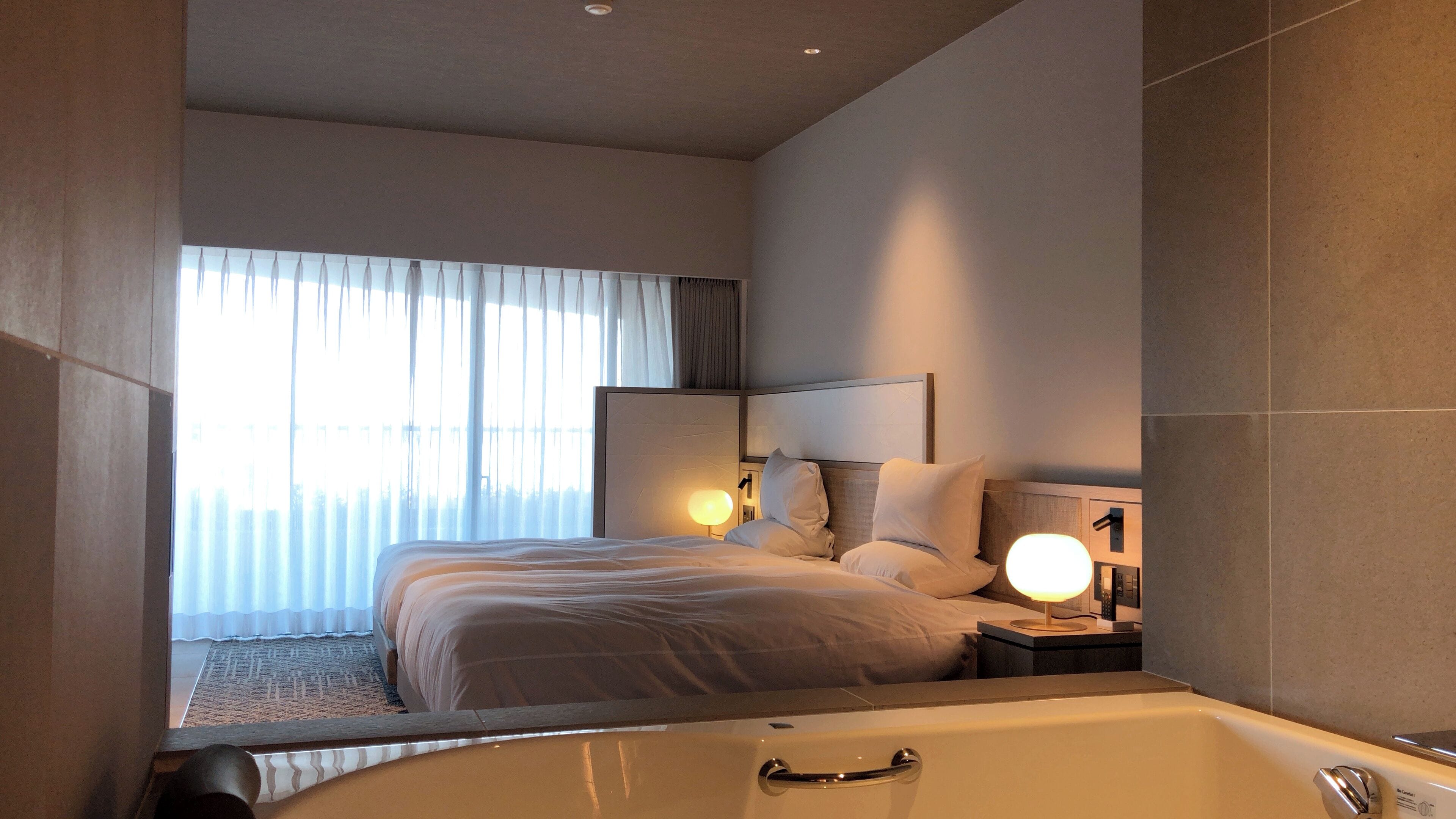 Ein modernes Hotelzimmer mit einem großen Bett, einem Nachttisch mit Lampe, einem großen Fenster mit Vorhängen und einem separaten Badezimmer mit Badewanne.