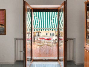 Una habitación con puerta corrediza de vidrio que da a un patio exterior con toldo a rayas, mobiliario blanco y suelo de baldosas.