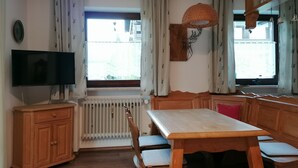 A dining area with a wooden table and chairs, a television on a stand, and a window with curtains. - Ferienwohnung 45 qm, für 2 Personen (Inzell)