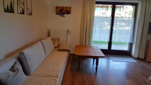 A room with a wooden floor, a sofa with cushions, a small table, and a window with curtains. - Ferienwohnung 45 qm, für 2 Personen (Inzell)
