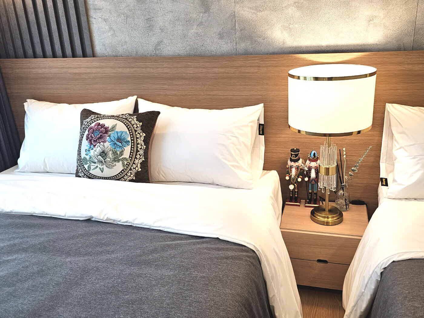 Una cama bien tendida con sábanas blancas, un cojín decorativo y una mesita de noche con una lámpara y figuritas de espantapájaros.