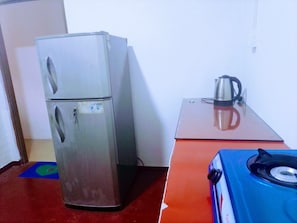 A compact kitchen with a refrigerator, a gas stove, and a kettle on a small table. - Volga Guest House (Weligama)