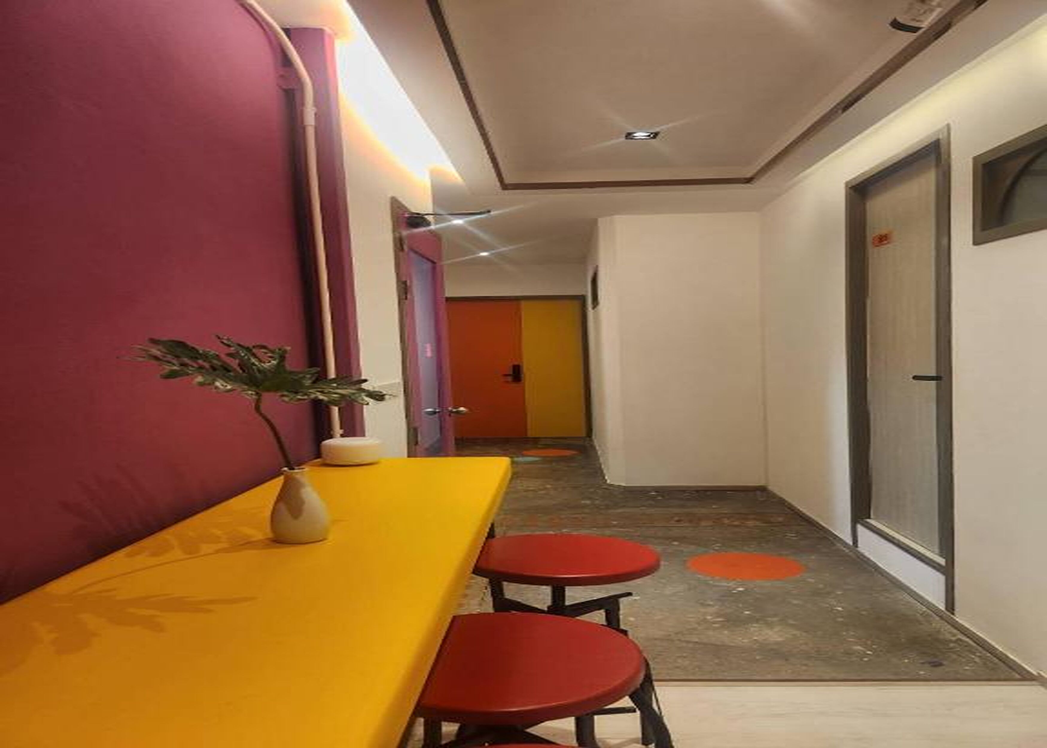 Un pasillo con una mesa amarilla y sillas rojas, una planta en maceta y una puerta con ventanal.