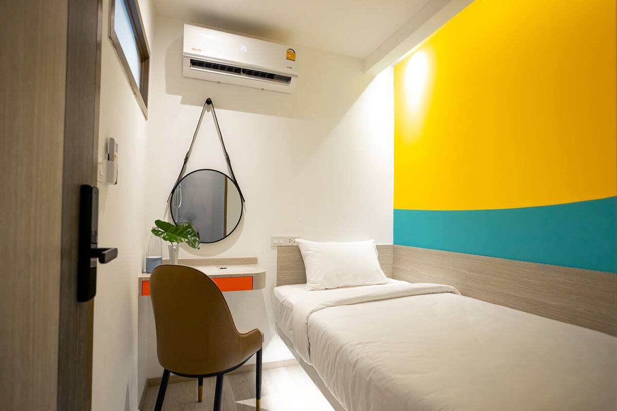 Quarto de hotel com uma cama, uma escrivaninha com cadeira, um espelho e um aparelho de ar condicionado.