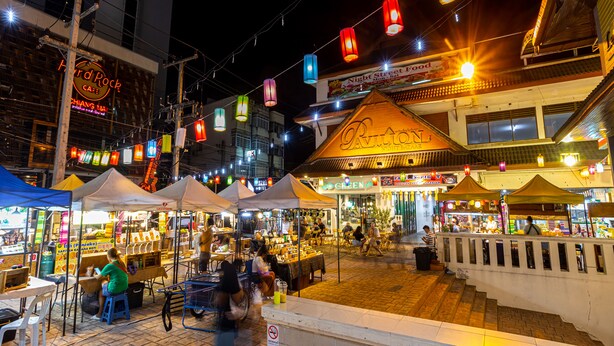 Un mercado callejero animado con varios puestos y carpas, un letrero destacado que dice 'Night Bazaar' y un letrero de 'Hard Rock Cafe'.
