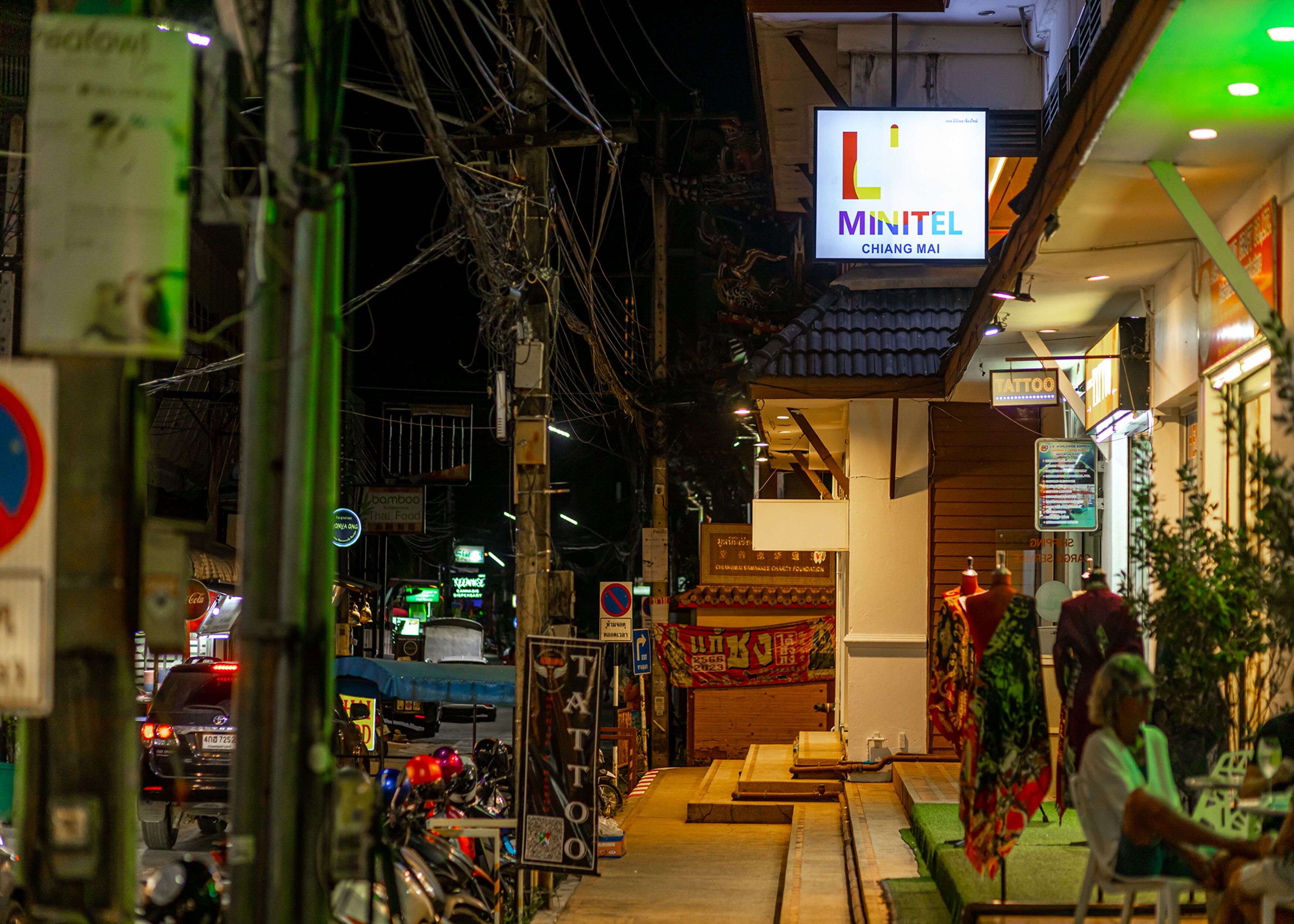 En smal gade med et skilt for 'L Minitel Chiang Mai' og en tydelig butiksfacade.