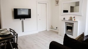A living room with a flat-screen TV, a fireplace, a dining area with a table and chairs, and a large mirror above the fireplace. - 4 Bed House, Sleeps 9, Free Parking (Stoke-on-Trent)