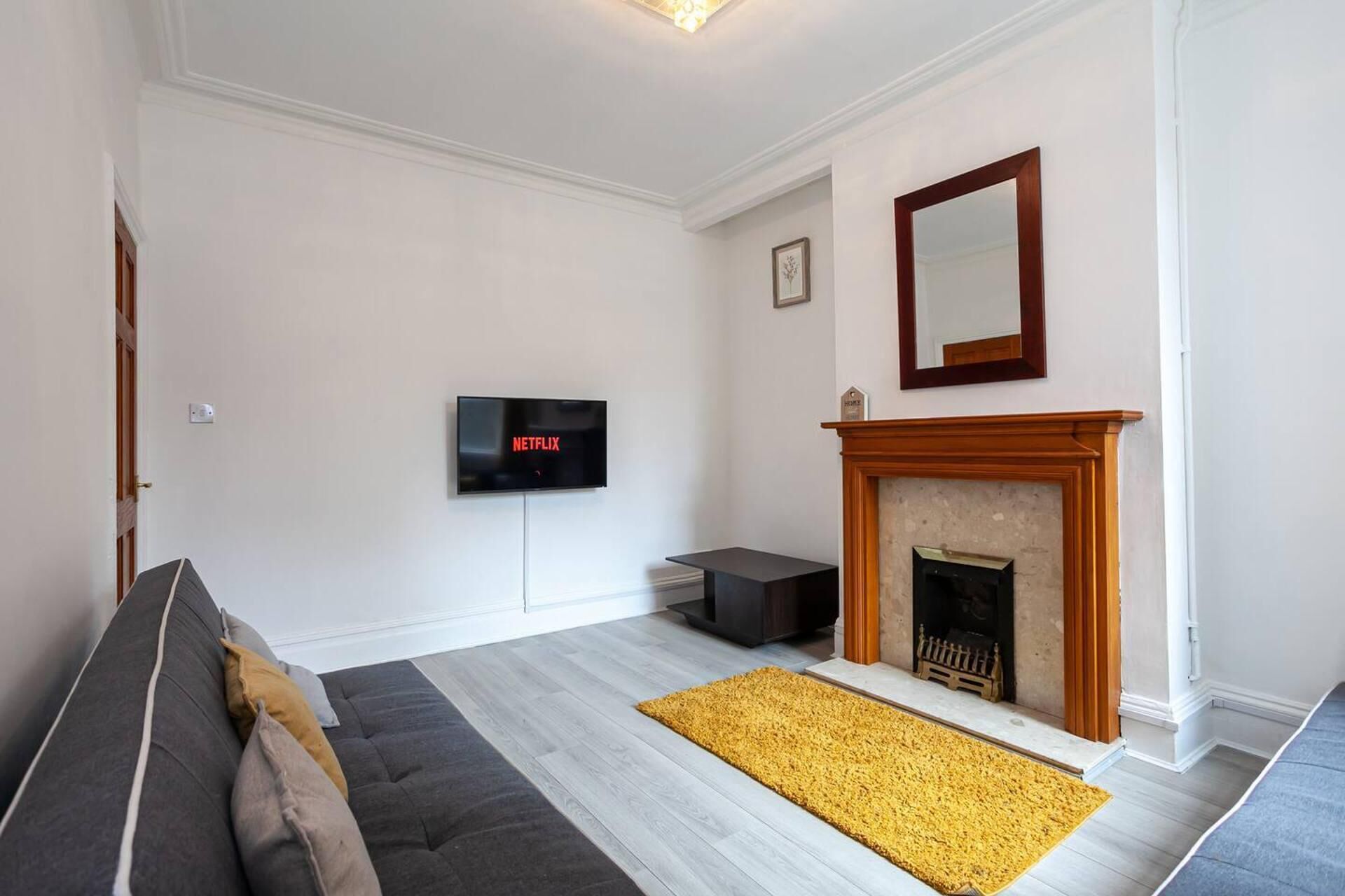 A living room with a sofa, a fireplace, a TV mounted on the wall, and a yellow rug.