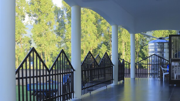 A covered walkway with white columns, black railings, and a view of trees. - Rainbow hotel-annex (Bundibugyo)