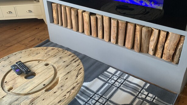 Una sala de estar moderna con un acuario integrado, troncos de madera como elemento decorativo en la pared, una mesa de centro redonda de madera y un televisor sobre un mueble blanco.