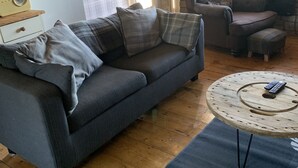 Una sala de estar acogedora con un sofá gris oscuro, un sillón escocés y una mesa redonda de centro con un control remoto.