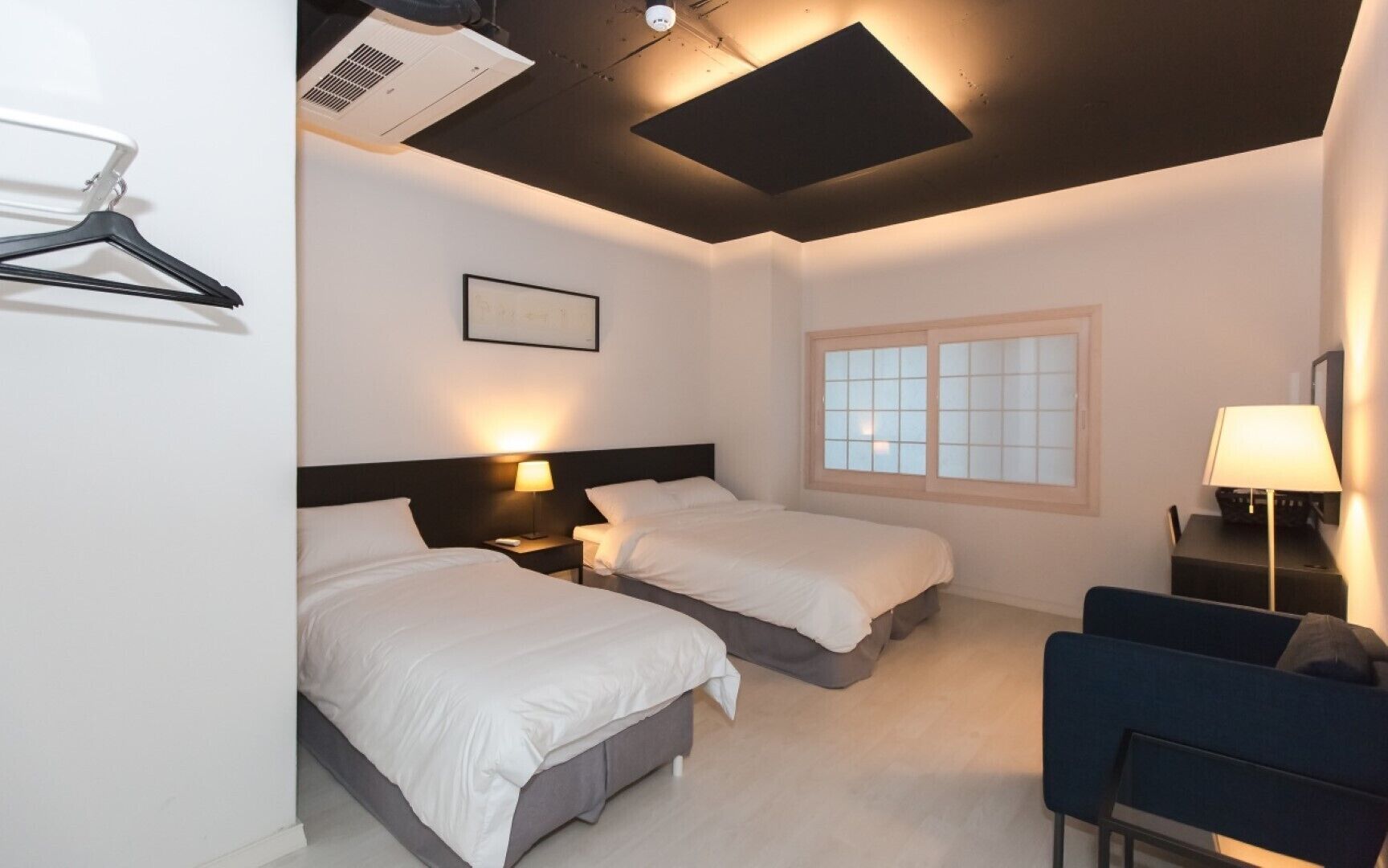 A hotel room with two beds, a desk, a chair, a window, and a wall-mounted air conditioner.
