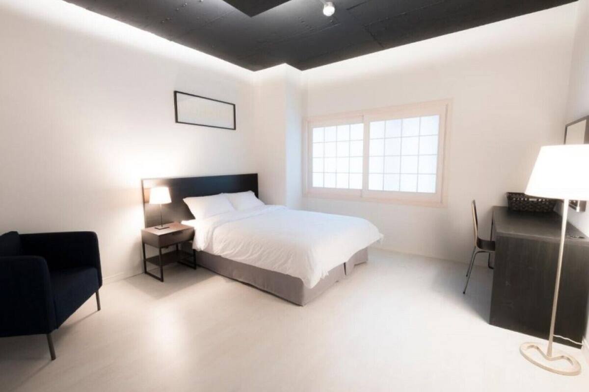 Ein modernes Schlafzimmer mit Bett, Nachttisch, Lampe, Stuhl, Schreibtisch und einem Fenster.