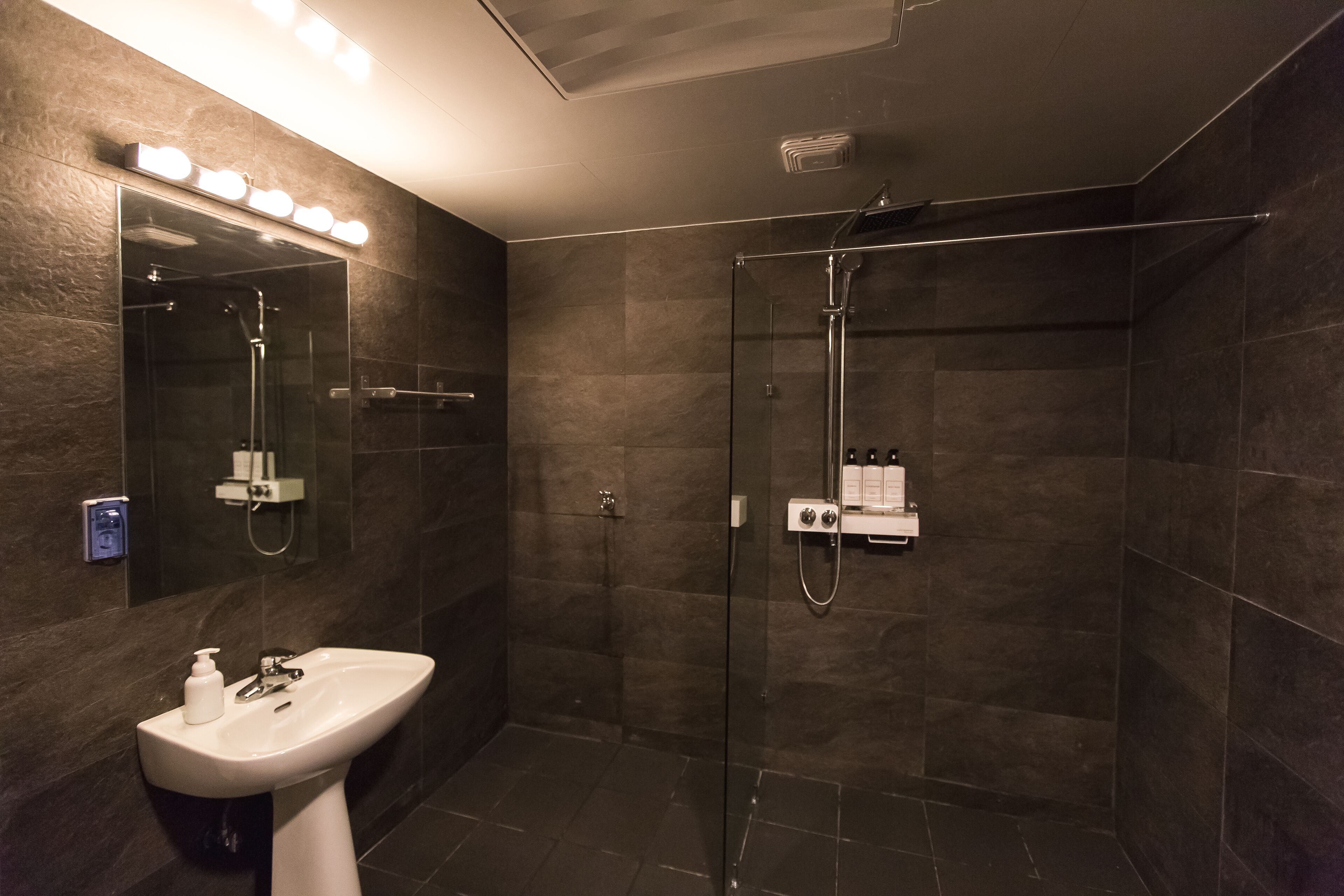 A modern bathroom with a glass-enclosed shower, a pedestal sink, and a mirror with lights above.