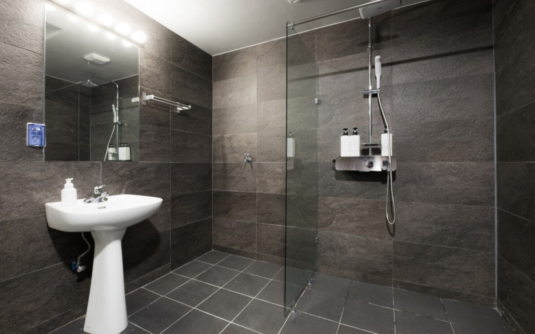 A modern bathroom with a glass-enclosed shower, a pedestal sink, and dark tiled walls.