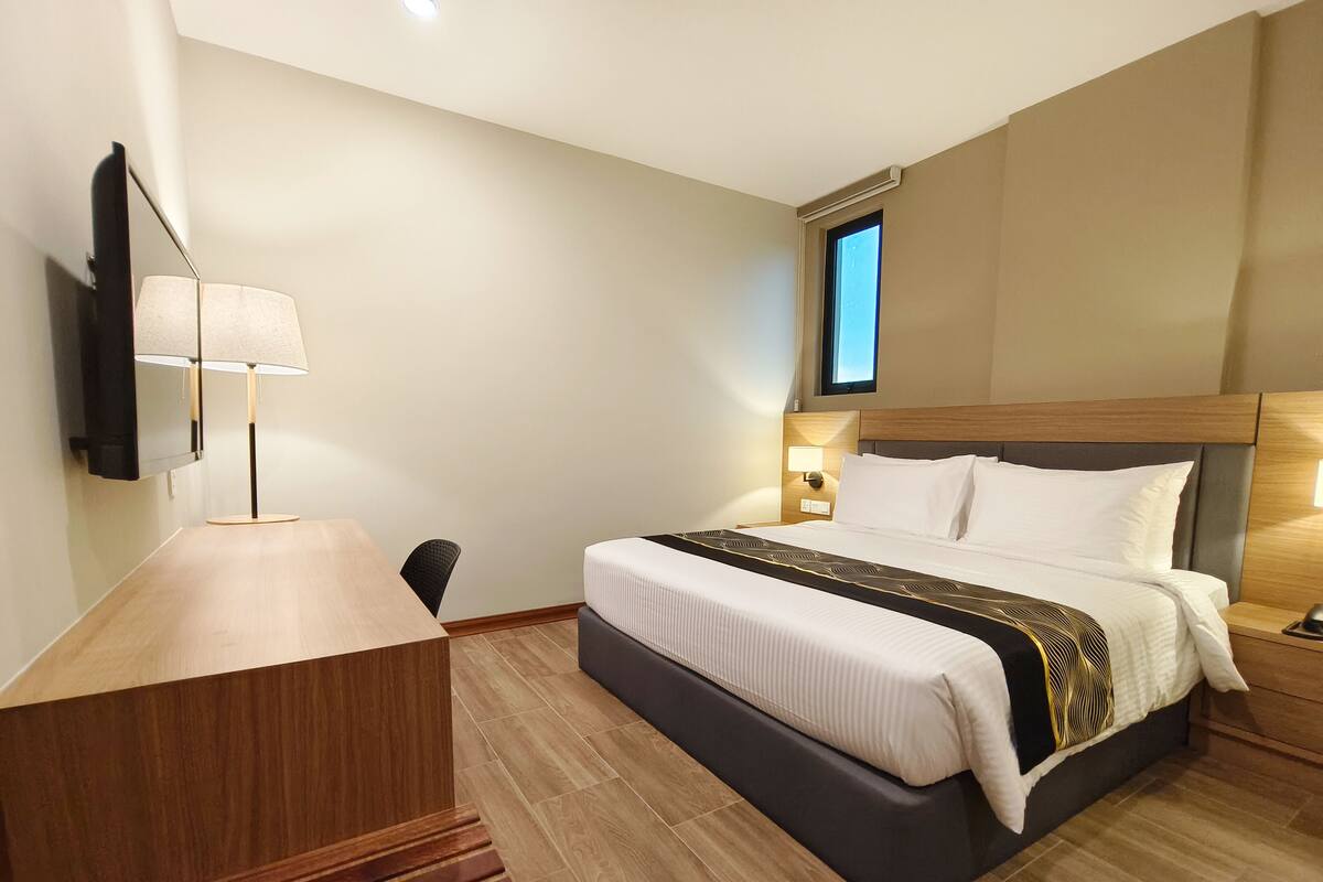 A hotel room with a wooden desk, a bed with a headboard, a nightstand with a lamp, and a window with a view.