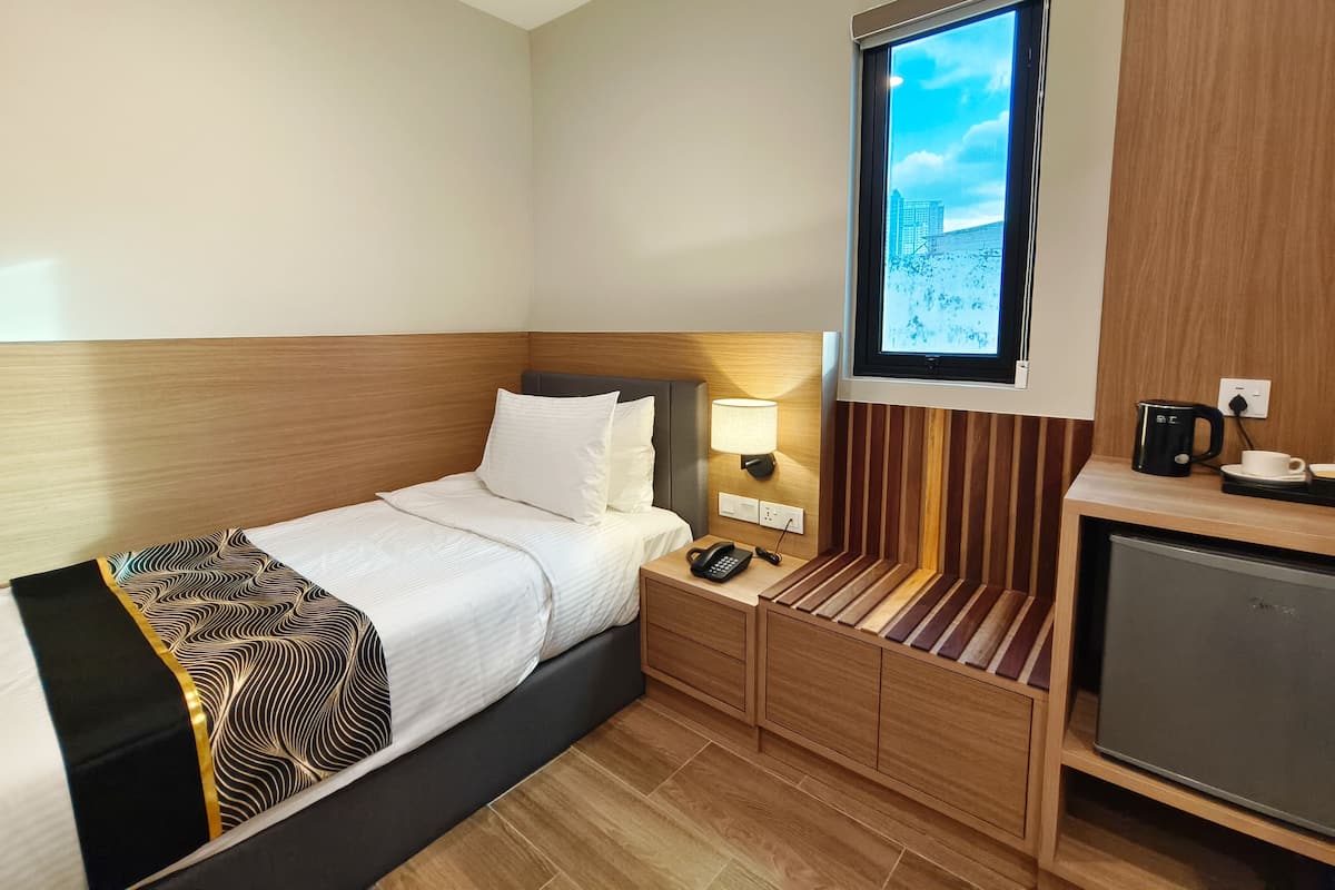 A hotel room with a bed, bedside table, lamp, and a window with a view of buildings.