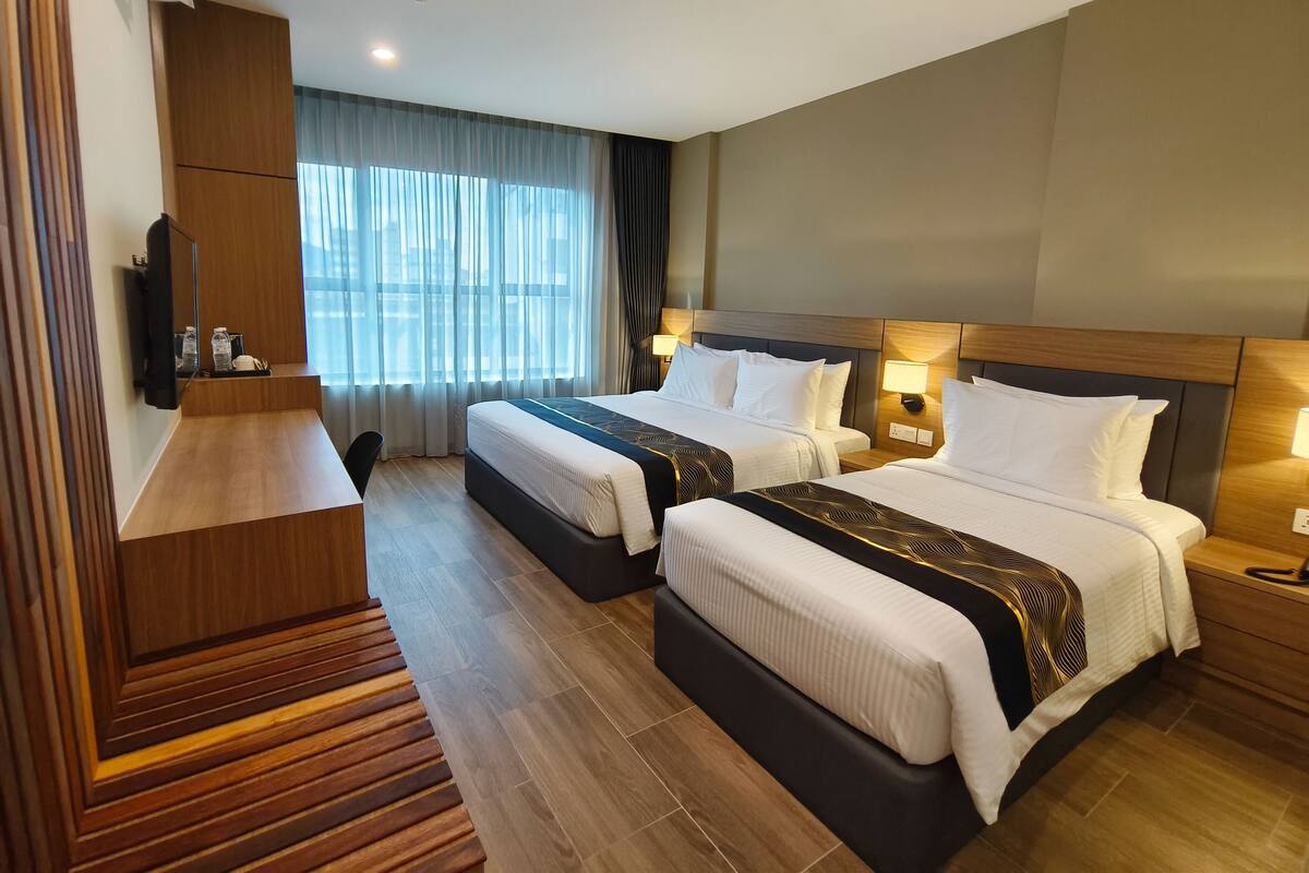 A hotel room with two beds, a wooden desk, a flat-screen TV, and a large window with curtains.