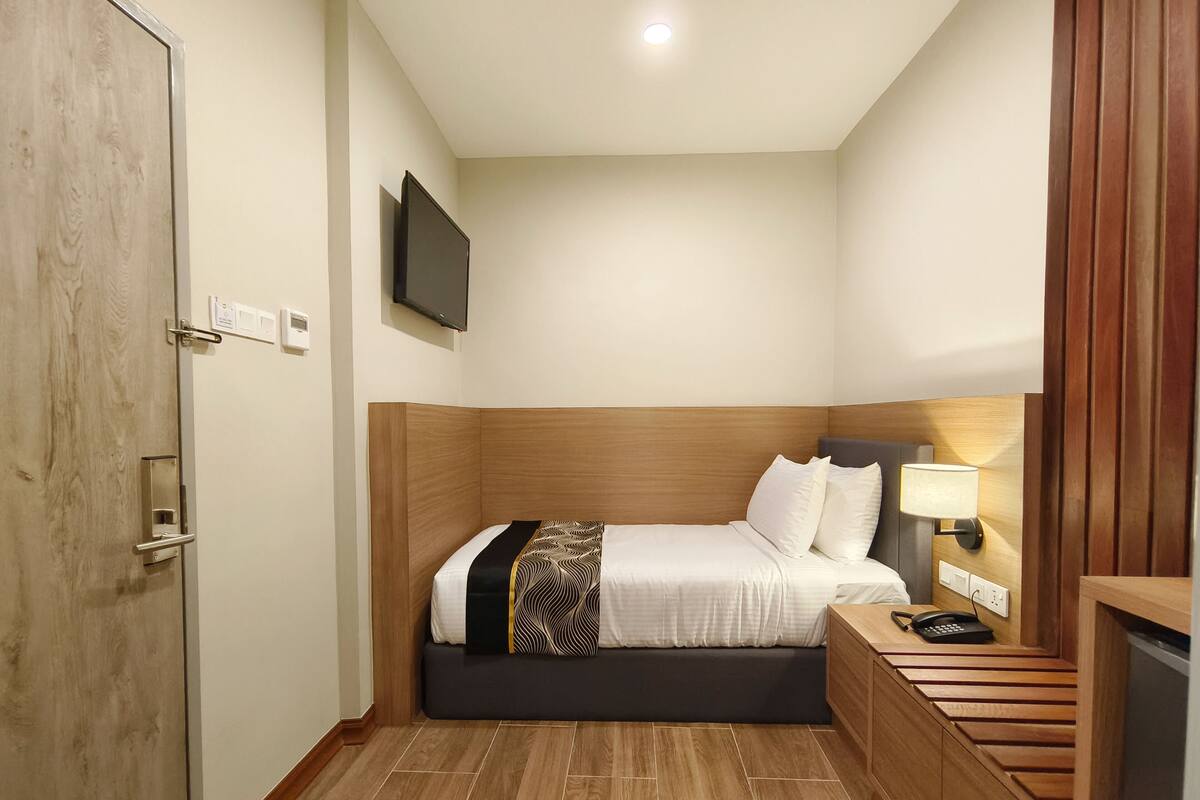 A hotel room with a wooden headboard, a bed with white bedding and a patterned throw, a bedside table with a lamp, and a TV mounted on the wall.