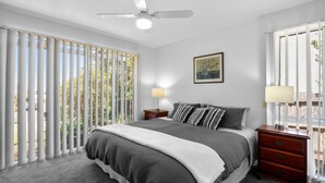 A bedroom with a bed, two bedside tables, a ceiling fan, a window with blinds, and a framed picture on the wall. - Three Bedroom House with Ocean Views (Torquay)