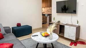 Un salon avec un canapé gris, un pouf bleu, une table basse en marbre sur laquelle se trouvent un panier de fruits et un vase, un téléviseur fixé au mur, et une cuisine en arrière-plan.