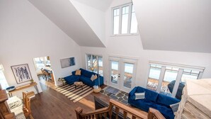 Un amplio salón con un sofá azul, piso de madera y vistas a la playa a través de grandes ventanales.