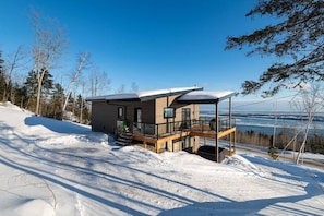 A modern cabin with a balcony, situated on a snowy slope with a view of a body of water.