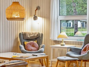 Ett mysigt rum med en väggpanel av trä, en grå fåtölj med en brun pläd, ett litet bord med en lampa och ett fönster med utsikt över träd.