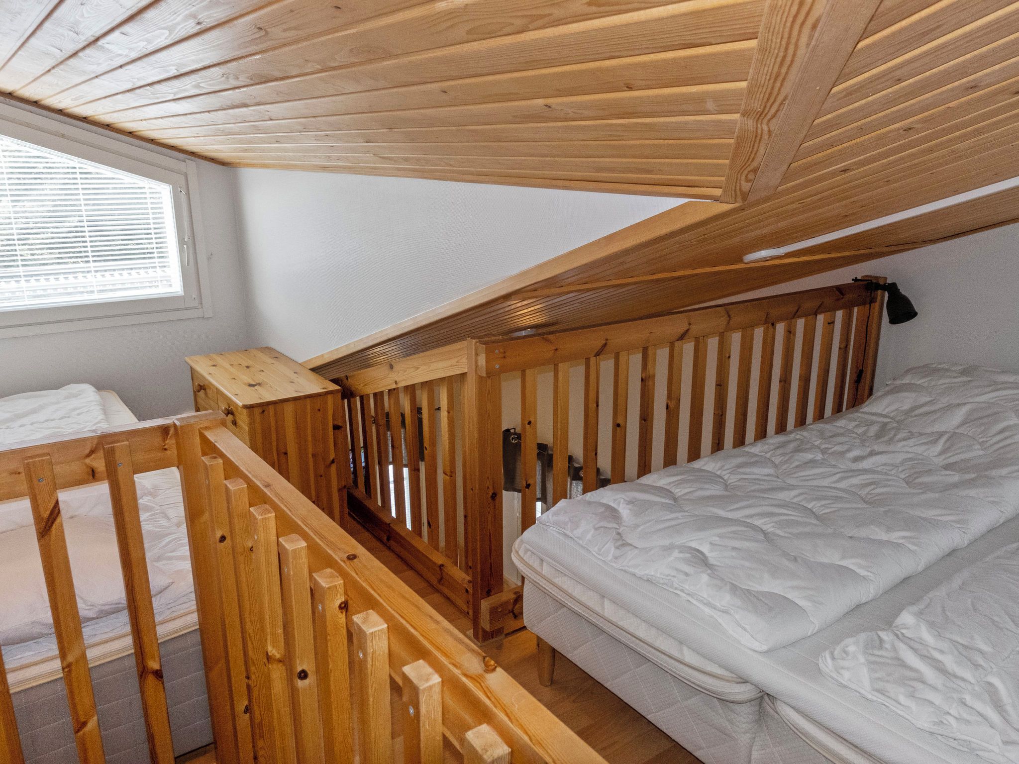Une chambre de dortoir avec un lit superposé, des rambardes en bois, une fenêtre avec des stores et un matelas blanc.