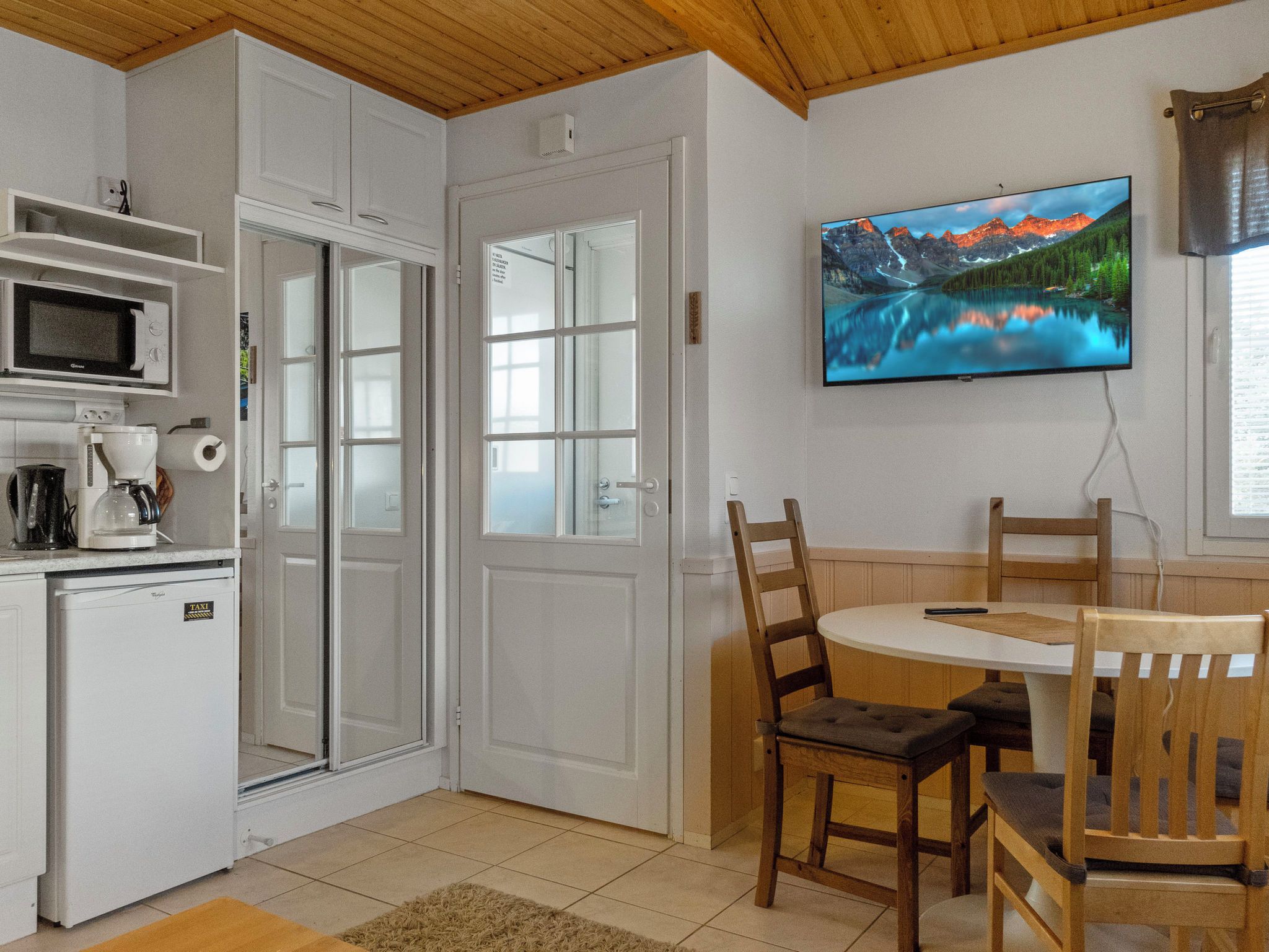 Ett kompakt kök med mikrovågsugn, kaffebryggare och ett litet kylskåp. Här finns också en matplats med ett bord och stolar, och en tv som är monterad på väggen och visar ett vackert landskap.
