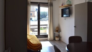 Een kamer met een bed, een tafel, een stoel, een televisie en uitzicht door een raam.