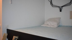 Een eenpersoonsbed met een witte matras en kussen, een houten stoel en een decoratief object aan de muur.