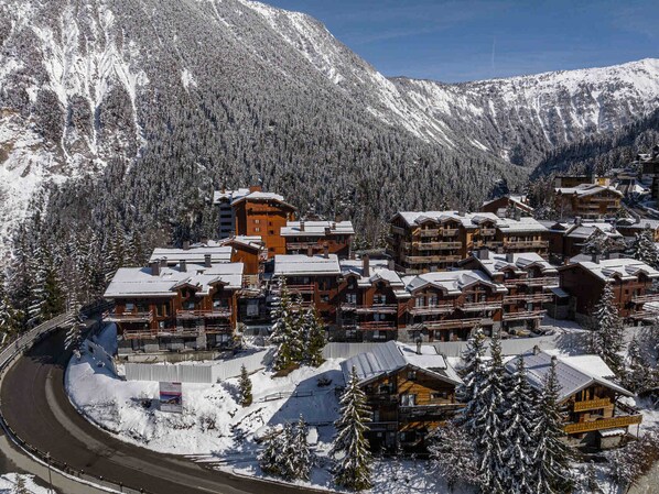 A snow-covered mountain resort with multiple wooden buildings, including lodges and a hotel, surrounded by dense forests and mountains.