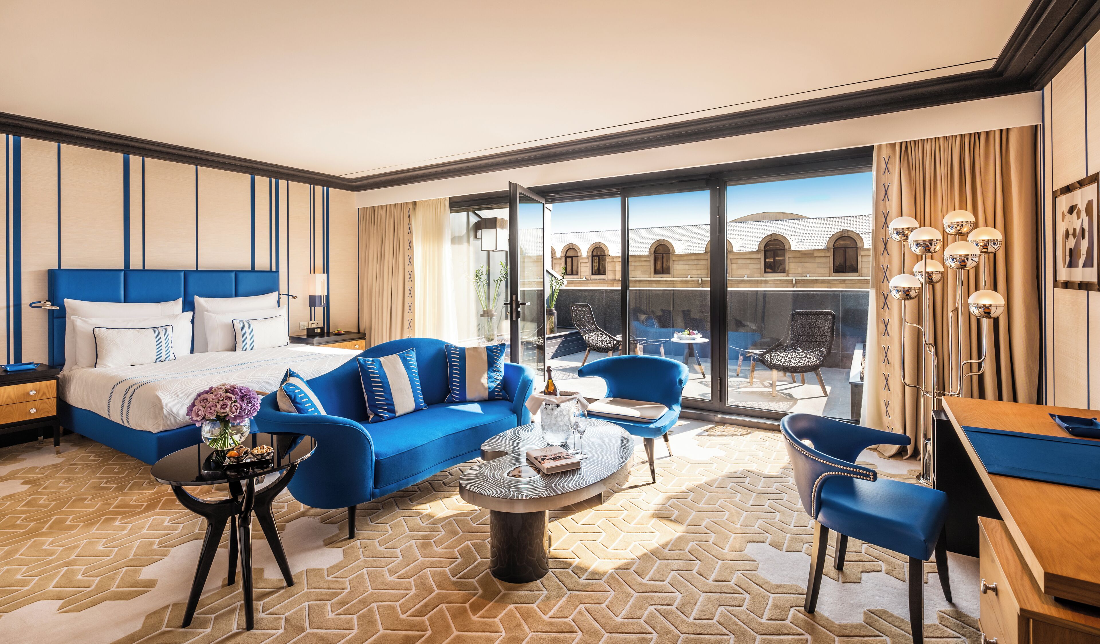 Ein modernes Hotelzimmer mit einem großen Bett, einem blauen Sofa, einem runden Couchtisch und einem Balkon mit Blick auf die Stadt.