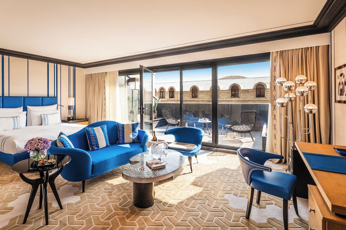 Ein modernes Hotelzimmer mit einem großen Bett, einem blauen Sofa, einem runden Couchtisch und einem Balkon mit Blick auf die Stadt.