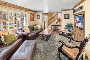 A spacious living room with a large window, a wooden staircase, and a television mounted on the wall.