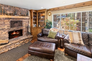 A cozy living room with a stone fireplace, leather sofas, a bookshelf, and a large window with a view of trees.