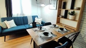 Ein modernes Wohnzimmer mit Essbereich, ausgestattet mit einem blauen Sofa, einem Holzesstisch für vier Personen und einem Bücherregal mit Pflanzen.