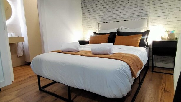 Ein modernes Schlafzimmer mit einer Ziegelwand, Holzboden, einem Bett mit weißer Bettwäsche und einem Nachttisch mit Lampe.