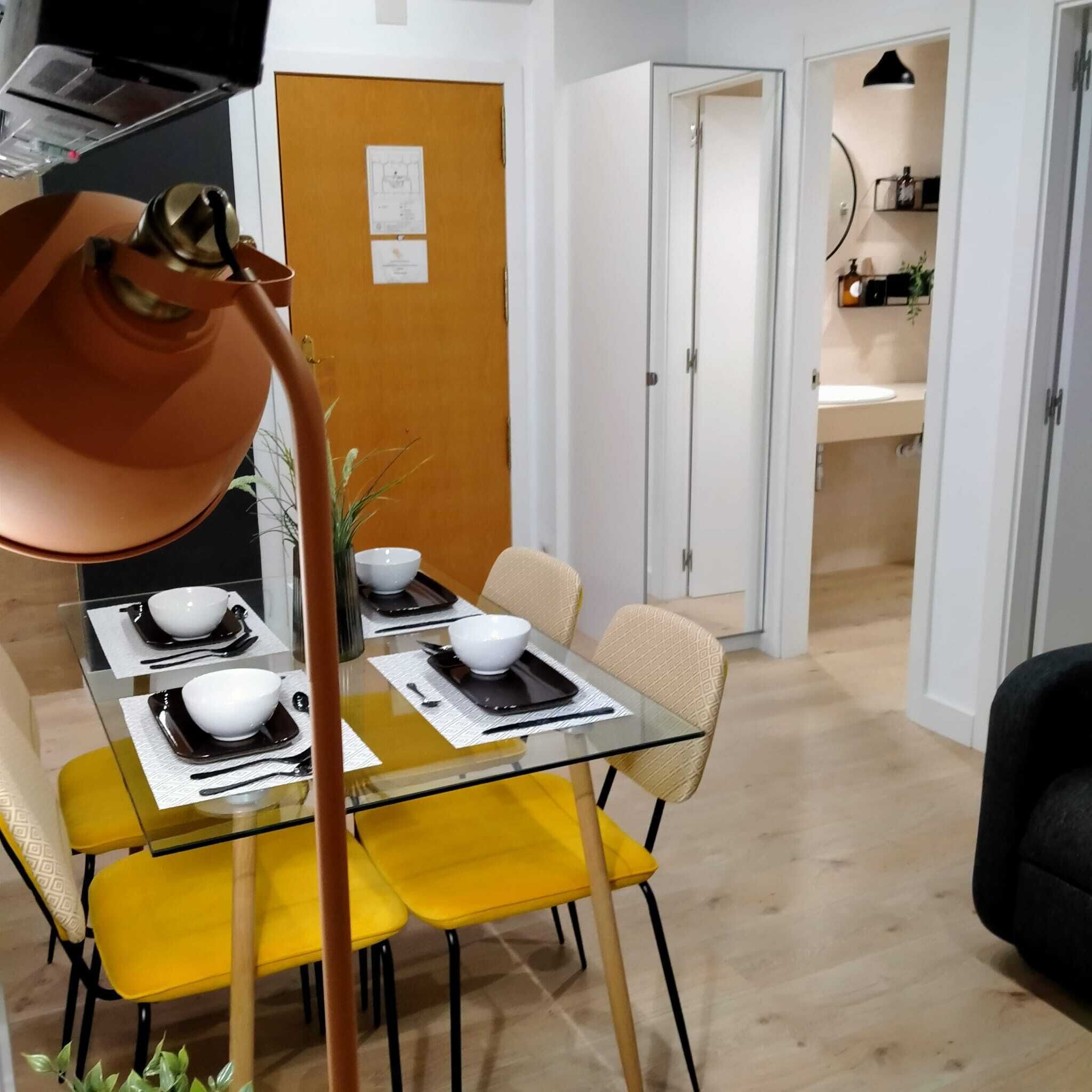 A dining area with a glass table set for four, yellow chairs, and a modern pendant light.