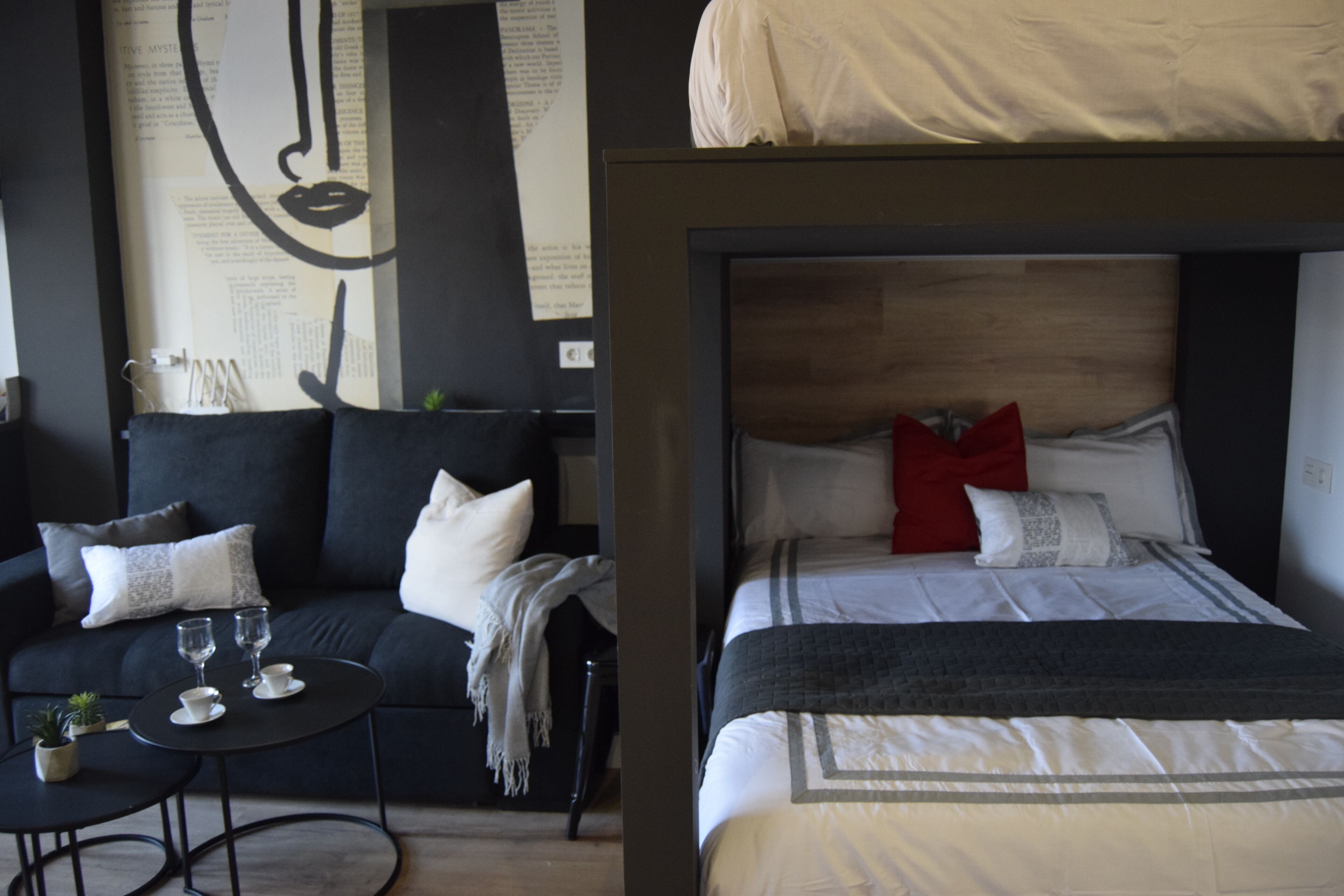 A modern hotel room with a black sofa, a small round table with two glasses and a cup, and a bed with a wooden headboard and multiple pillows.