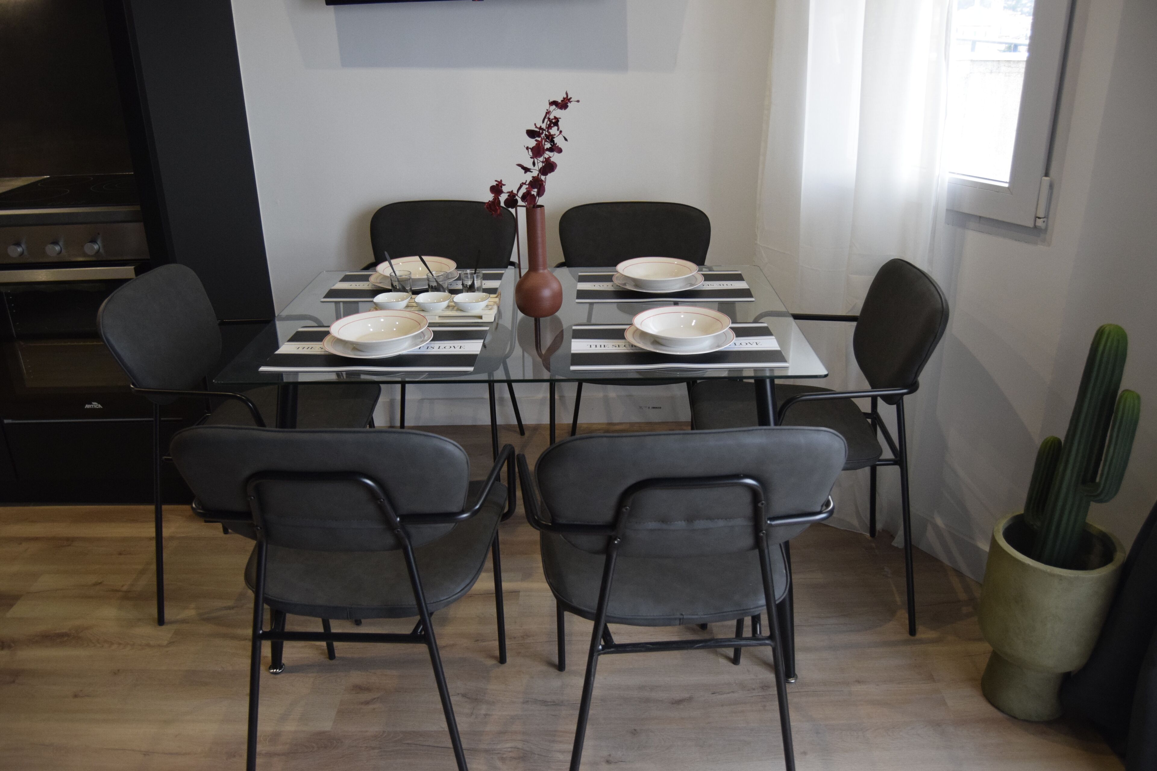 Een eethoek met een tafel voor vier personen, omringd door stoelen, een bloevas en een cactus.