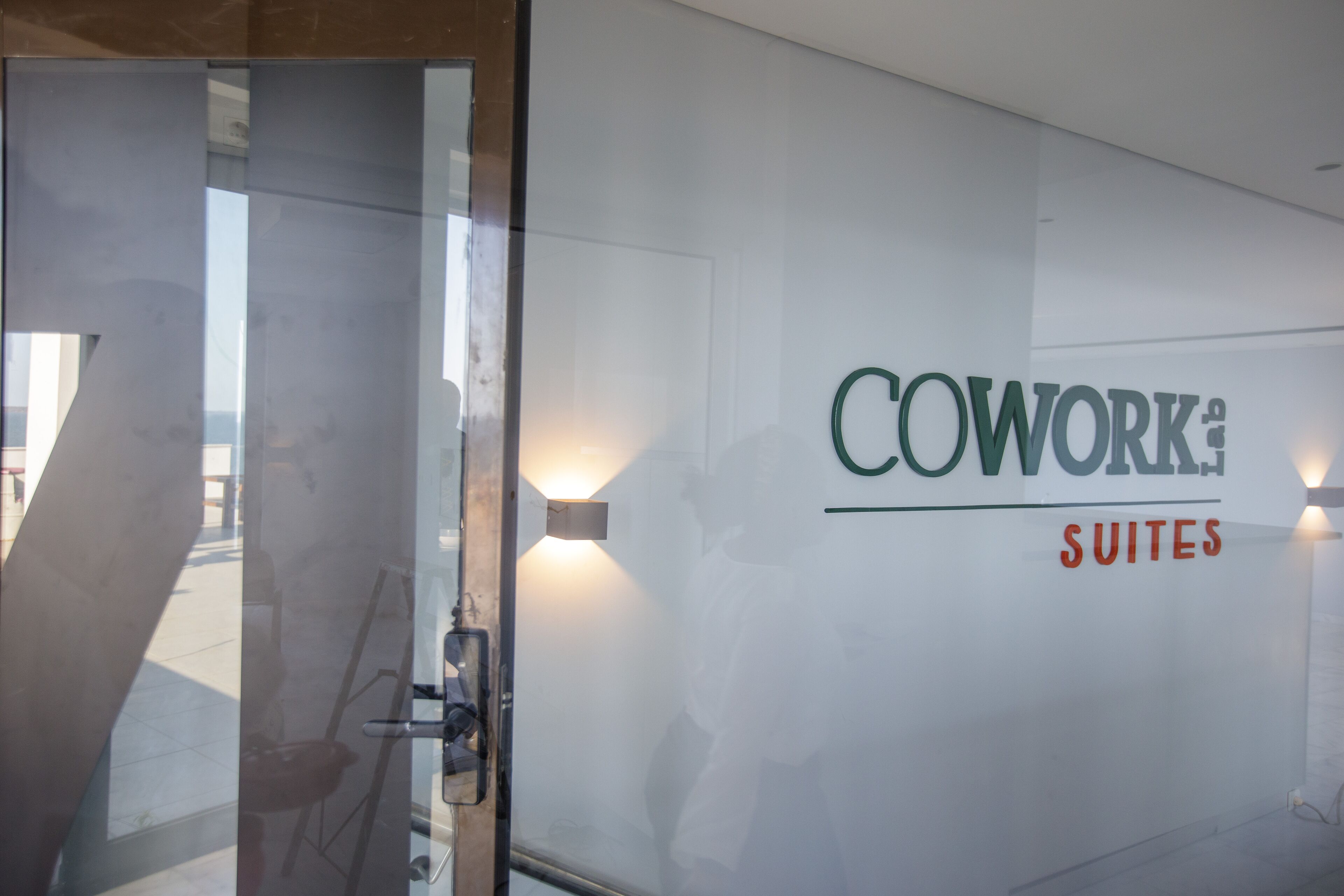 A glass door with a reflection of a person and a chair, and a wall with the text "COWORK: SUITES".