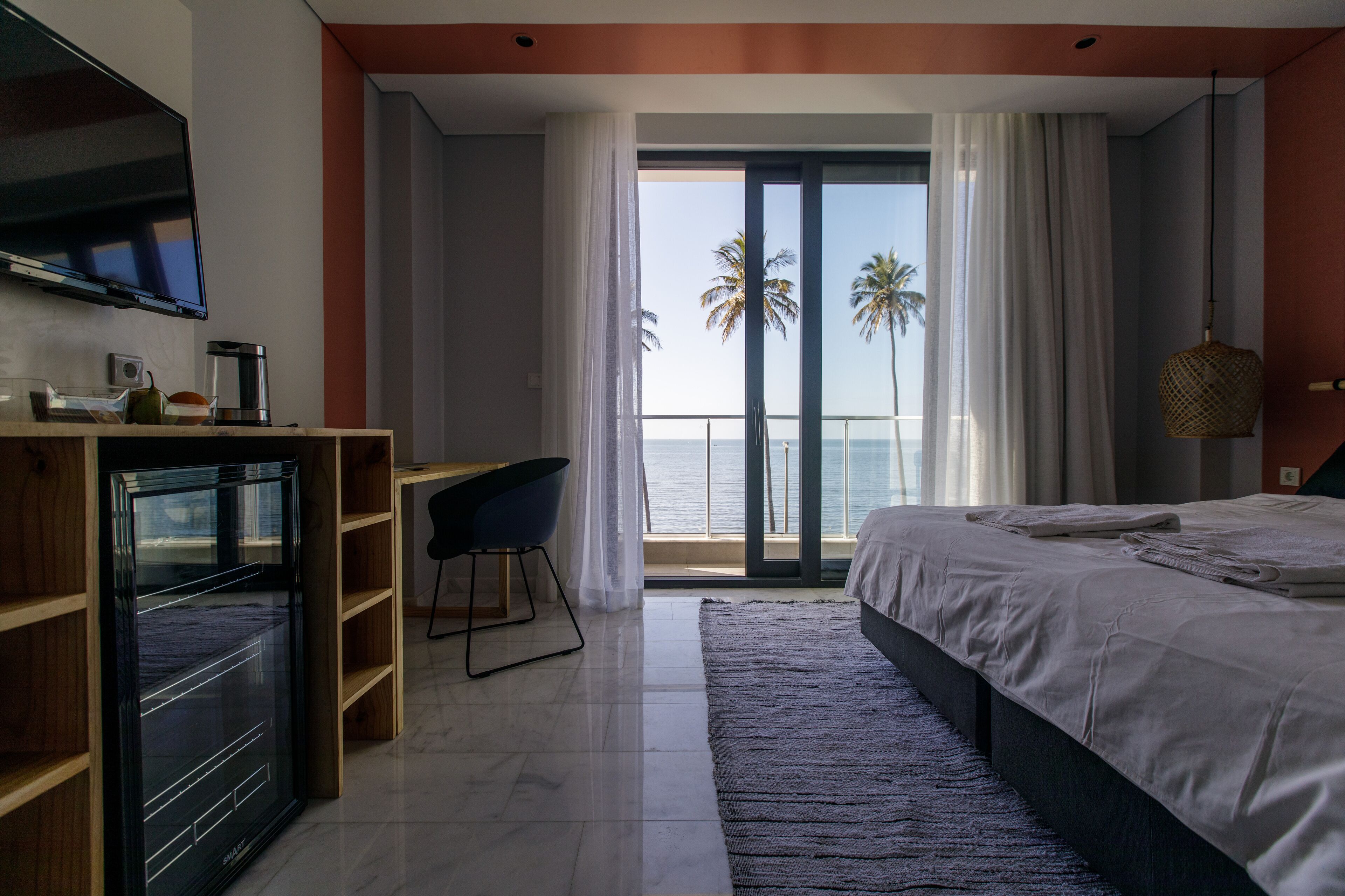 Un dormitorio moderno con una cama grande, un escritorio y un televisor. Hay un balcón con vistas a palmeras y al mar.