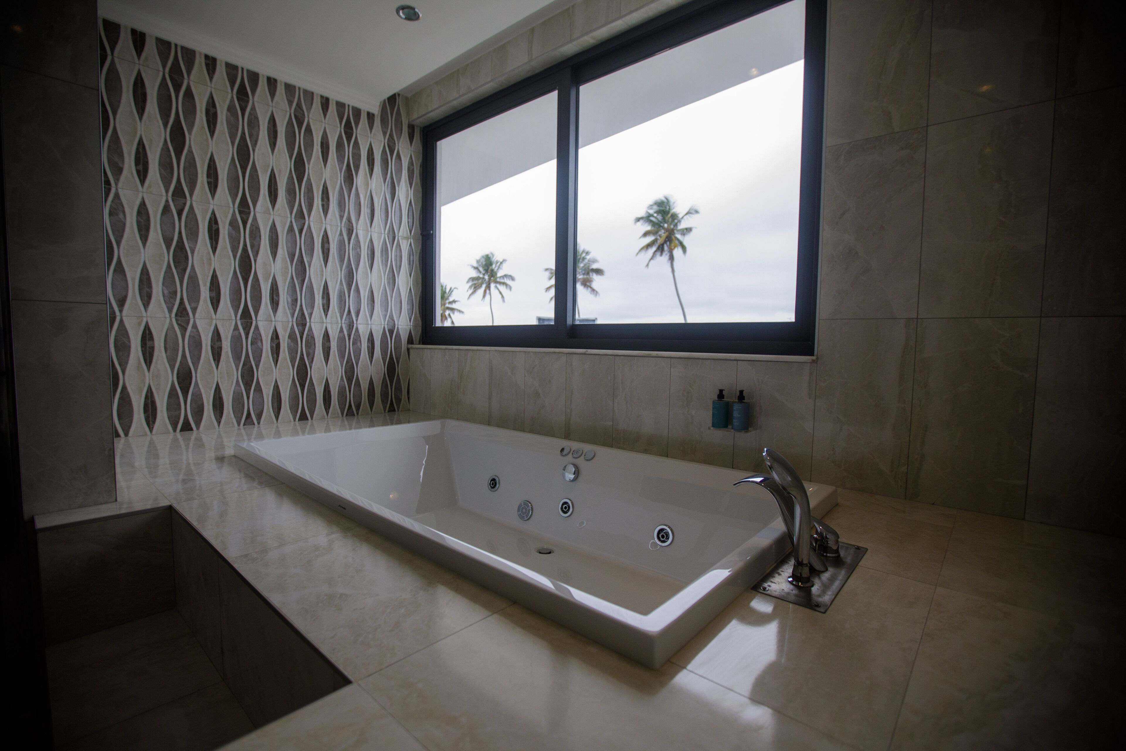 Un baño moderno con una bañera grande, una ventana que ofrece vistas a palmeras y un diseño decorativo en la pared.