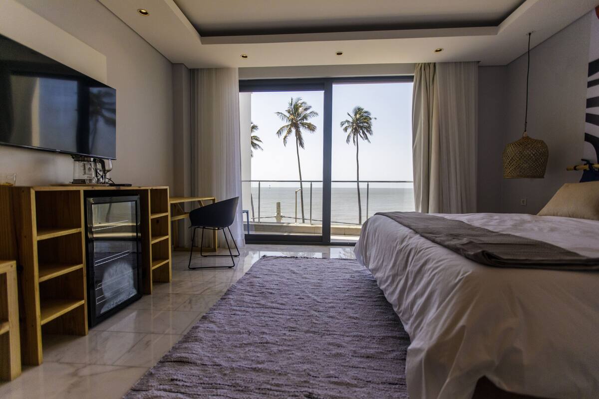 A modern hotel room with a large bed, a flat-screen TV, a desk with a chair, and a view of the ocean with palm trees.