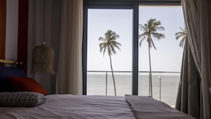 A bedroom with a large bed, a hanging light fixture, and a view of the sea with palm trees. - Cowork Lab Suites (Maputo)