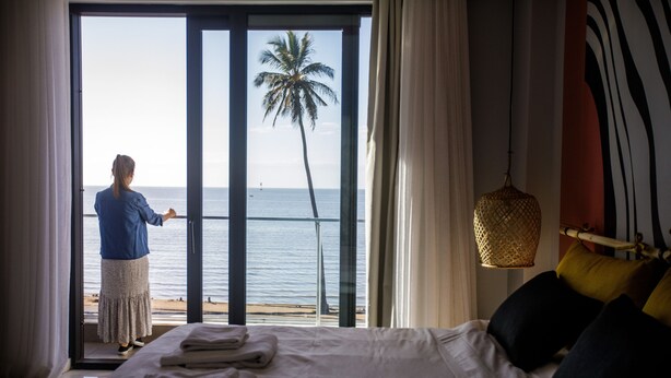 A hotel room with a bed, a view of the sea, and a person standing by the window. - Cowork Lab Suites (Maputo)