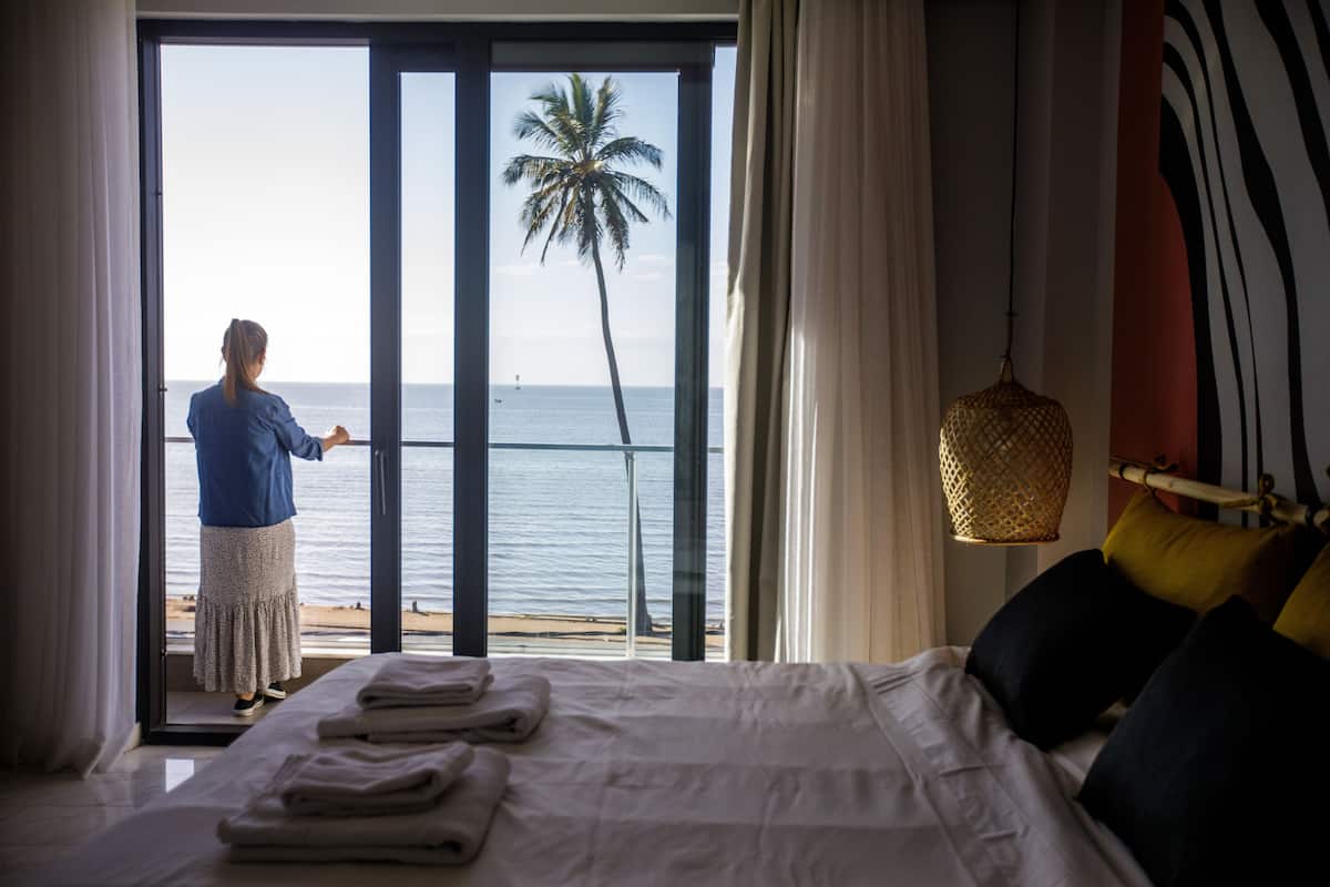 A hotel room with a bed, a view of the sea, and a person standing by the window.