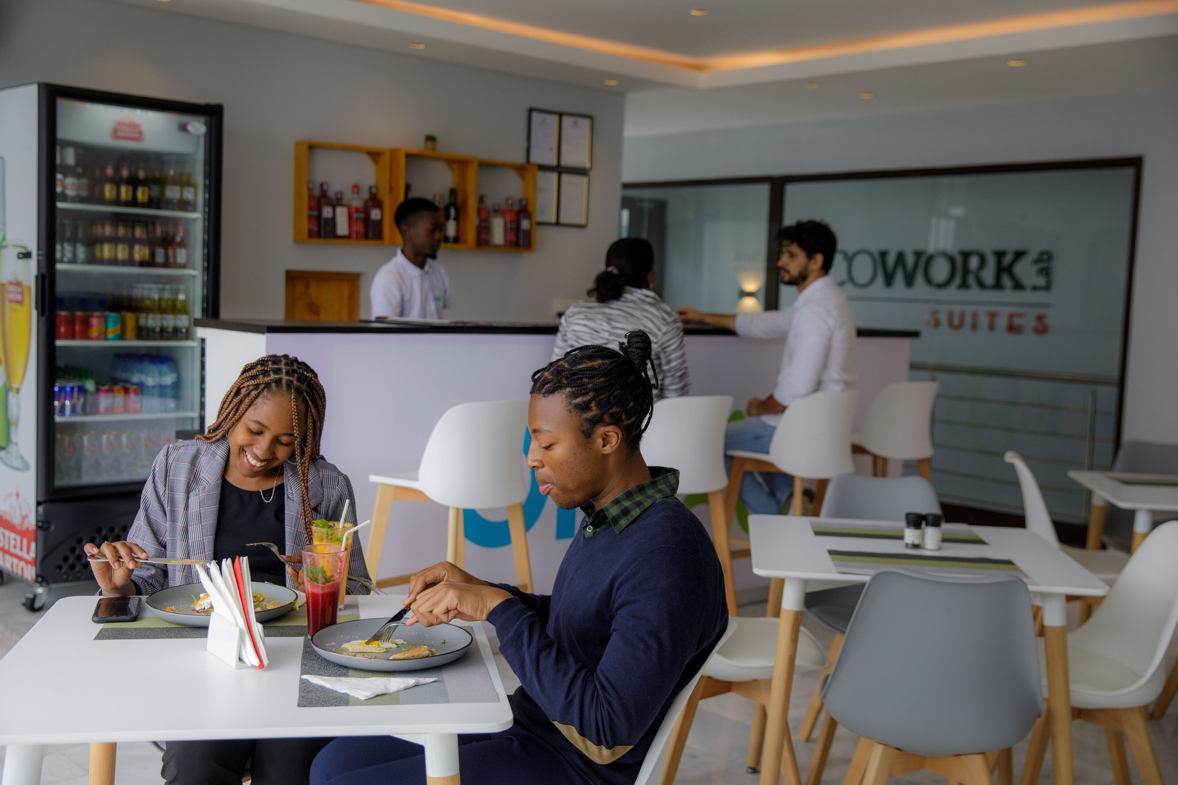 Un espace de restauration moderne avec des tables et des chaises blanches, un réfrigérateur rempli de boissons et une enseigne bien visible indiquant « Cowork4 Suites ».